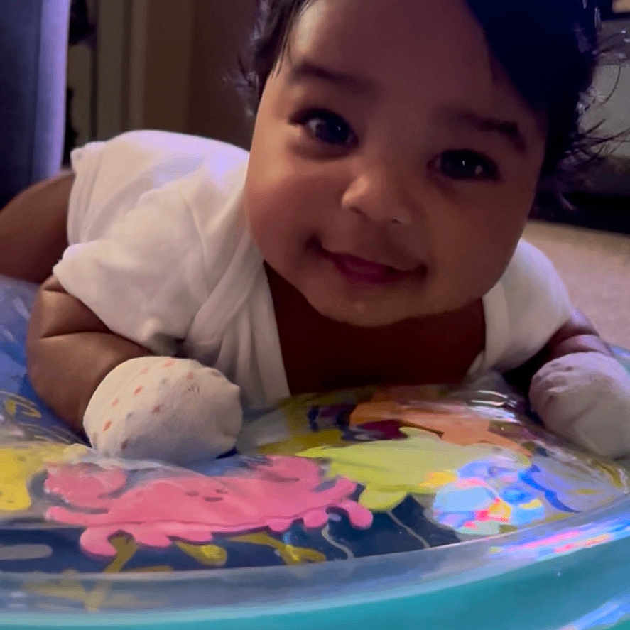 Kaeliah joined the competition — help win amazing prizes! baby, carpet, child, closeup, colorful, cute, dark_hair, face, happy, indoor, infant, inflatable_ring, lying_down, mittens, person, portrait, smiling, tummy_time, white_clothing, young