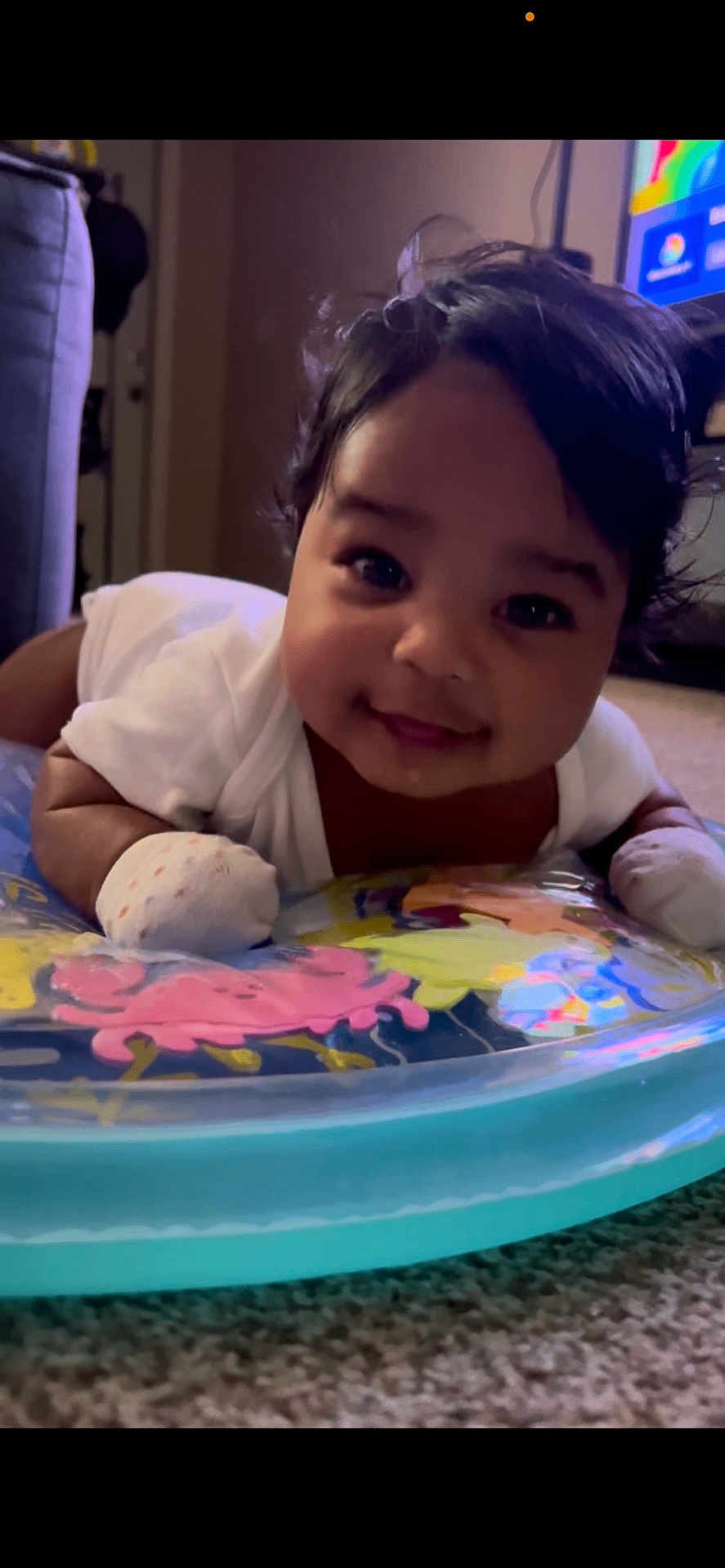 Kaeliah joined the competition — help win amazing prizes! baby, infant, smiling, face, dark_hair, mittens, white_clothing, inflatable_ring, colorful, carpet, indoor, child, cute, portrait, lying_down, tummy_time, happy, person, young, closeup
