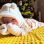 baby, infant, mittens, hood, bear_ears, yellow_blanket, christmas_tree, tinsel, ornaments, holiday, cozy, cute, indoors, sleepwear, patterned_clothing, soft_texture, child, smiling, portrait, festive
