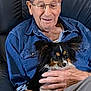 elderly_man, glasses, blue_shirt, dog, small_dog, black_dog, white_dog, fluffy_dog, sitting, couch, leather_couch, hand, smiling, indoors, comfortable, companionship, pet, casual_clothing, relaxed, portrait