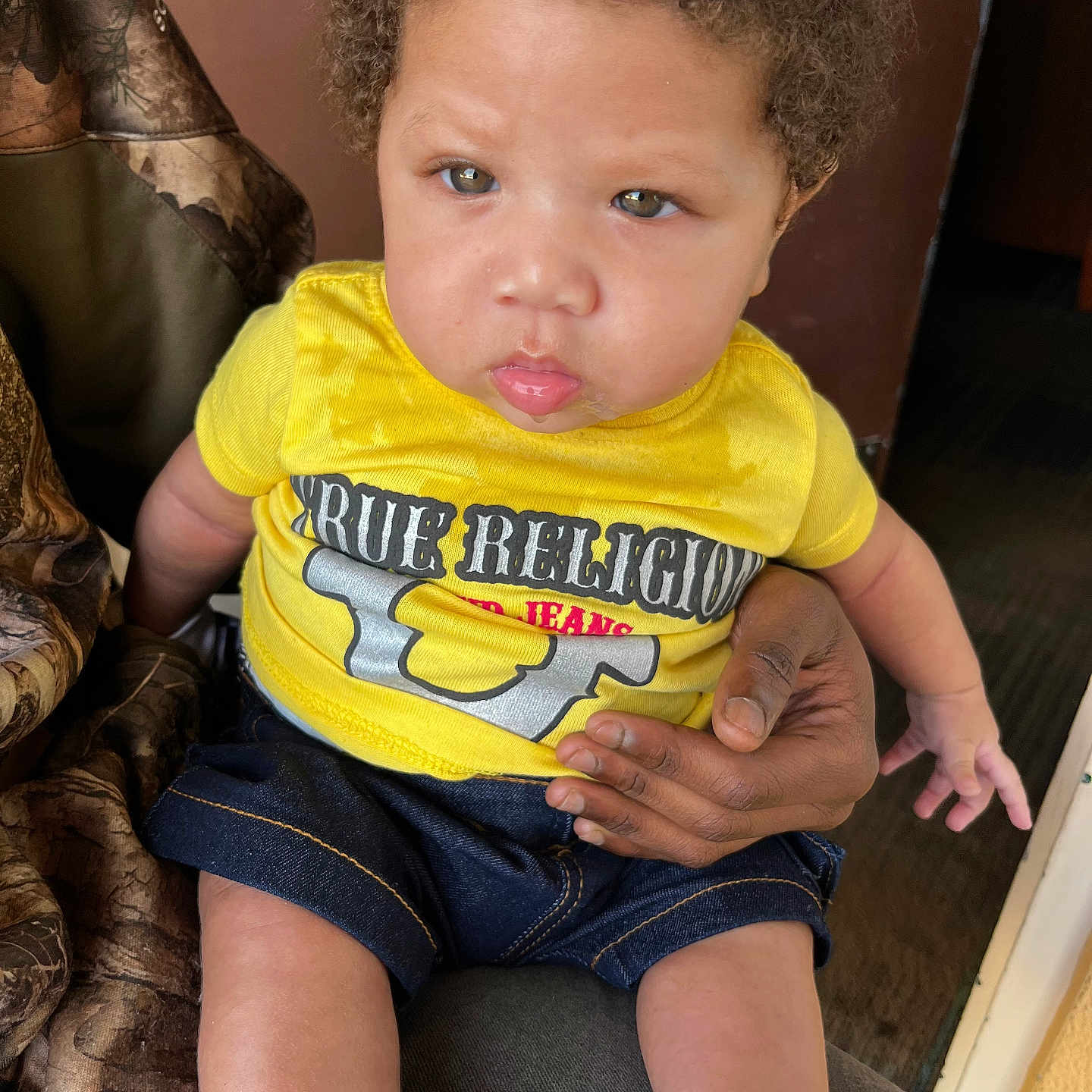 Raii is registered to the contest to win money with this photo: adult_hand, baby, black_shoes, casual, child, clothing, curly_hair, denim_shorts, expression, face, footwear, hand, holding, indoor, person, portrait, seated, skin, toddler, yellow_shirt