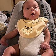 Braylon is registered to the contest to win money with this photo: baby, infant, bib, yellow, clothing, person, lap, sweatpants, text_on_clothing, hand, foot, sock, indoors, room, door, fabric, smile, cute, resting, expression