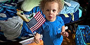 Carter is registered to the contest to win money with this photo: child, toddler, curly_hair, american_flag, blue_shirt, barefoot, wooden_floor, bed, blanket, toy, indoor, home, floor, person, blue_jeans, expression, standing, room, casual_clothing, light