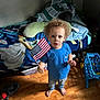 child, toddler, curly_hair, american_flag, blue_shirt, barefoot, wooden_floor, bed, blanket, toy, indoor, home, floor, person, blue_jeans, expression, standing, room, casual_clothing, light