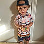 child, boy, barefoot, cap, backward_cap, curly_hair, toy, colorful_toy, racing_outfit, shorts, tshirt, indoor, tile_floor, shadow, door, wall, standing, looking, casual_clothing, cute