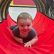 Damien is registered to the contest to win money with this photo: baby, child, smiling, crawling, play_tunnel, red, indoor, gym, floor, toddler, happy, cute, person, play, fabric, head, hand, clothing, young, fun