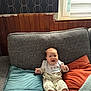 baby, smiling, couch, pillow, window, indoor, child, happy, bib, clothing, patterned_wallpaper, wood_paneling, natural_light, infant, sitting, face, person, furniture, blanket, home