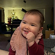 Romy participe au concours pour gagner de l'argent avec cette photo : baby, child, smiling, indoor, living_room, christmas_tree, bib, clothing, chewing, string, furniture, blurred_background, happy, portrait, infant, cute, person, warm_lighting, holiday_decor, cozy