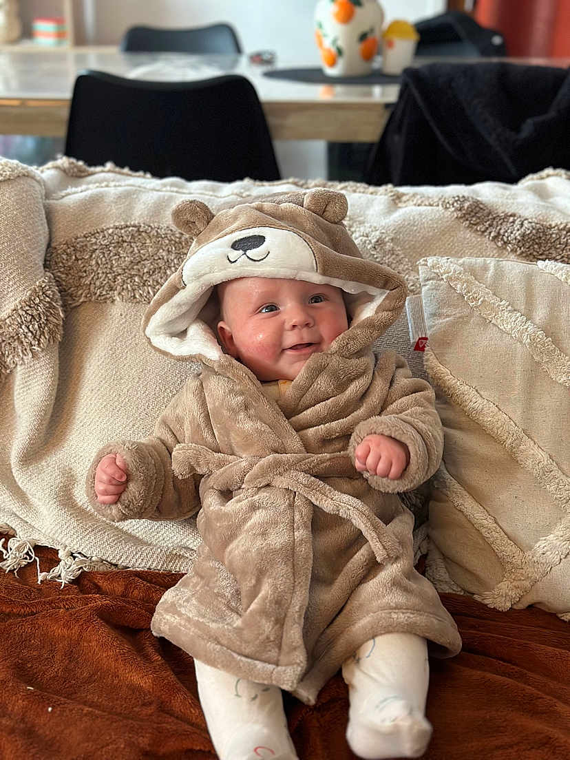 Eden a rejoint le concours — aidez-le/la à gagner de superbes lots ! baby, smiling, robe, bear_hood, couch, pillows, blanket, indoor, infant, child, furniture, soft_texture, cozy, happy, seated, face, headwear, living_room, warm_clothing, cute
