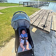 Casey is registered to the contest to win money with this photo: baby, stroller, outdoor, lake, water, grass, picnic_table, sunny, daylight, smiling, bib, infant, park, wood, bench, footwear, concrete, nature, child, happy
