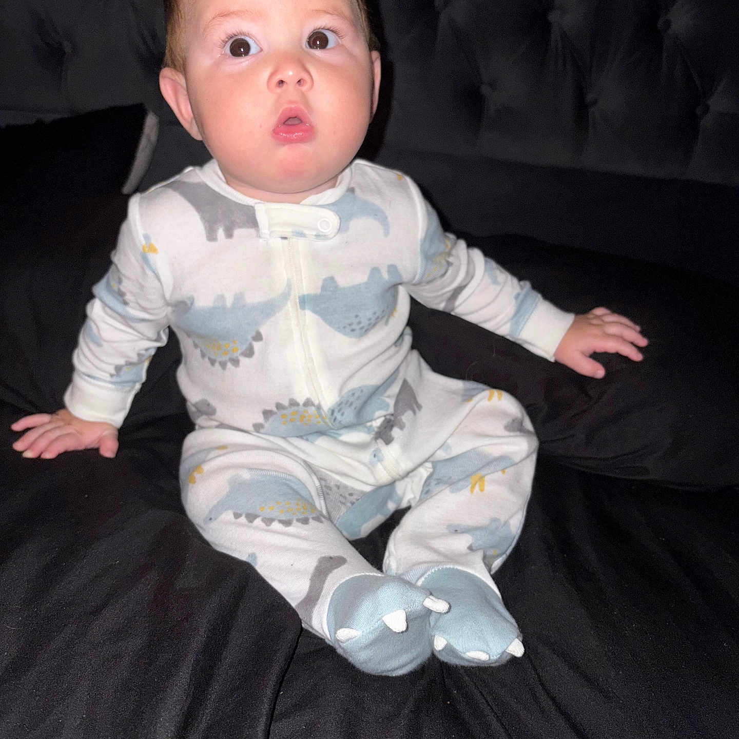 Austhin Gabriel joined the competition — help win amazing prizes! baby, bed, black_bedspread, child, cozy, cute, dinosaur_pattern, face, footed_pajamas, furniture, head, indoor, infant, pajamas, person, sitting, sleepwear, soft_lighting, surprised_expression, young_child
