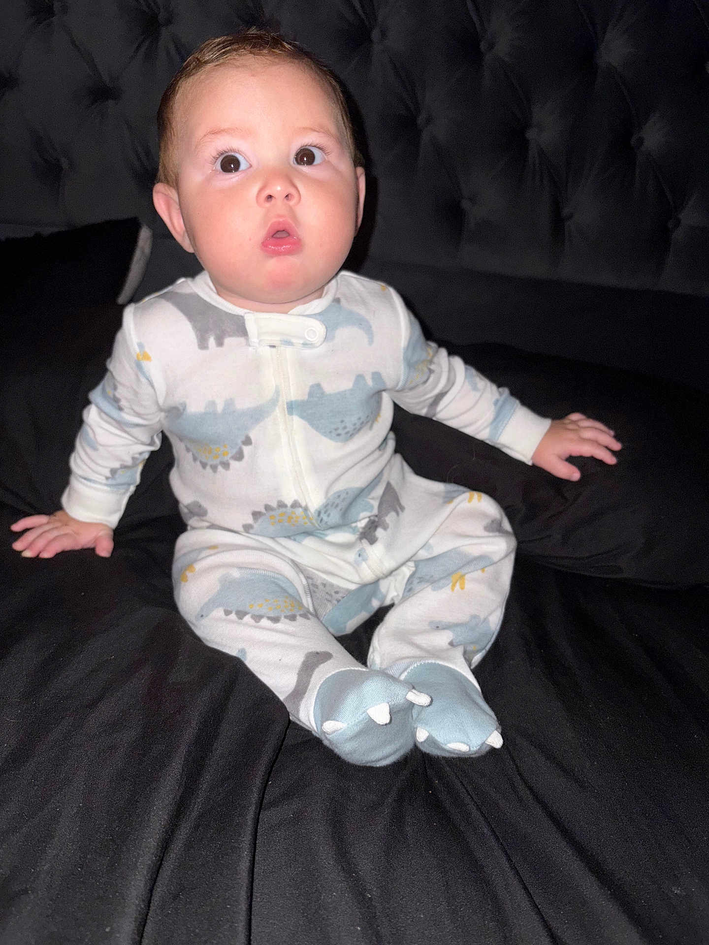 Austhin Gabriel joined the competition — help win amazing prizes! baby, child, pajamas, dinosaur_pattern, bed, black_bedspread, sitting, cute, surprised_expression, indoor, infant, person, sleepwear, footed_pajamas, head, face, furniture, soft_lighting, cozy, young_child