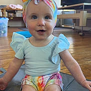 Aria is registered to the contest to win money with this photo: baby, blue_eyes, casual_clothes, child, colorful_clothing, curious_expression, cute, floor, headwear, headwrap, indoor, infant, living_room, mat, person, shorts, sitting, smiling, toddler, wooden_floor