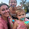 child, toddler, adult, face_paint, blue_frosting, cupcake, outdoor, grass, smiling, playful, tongue_out, bunny_ears, family, summer, trees, casual_clothing, happy, celebration, fun, portrait