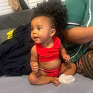 Kali joined the competition — help win amazing prizes! baby, child, curly_hair, red_clothing, sitting, couch, indoor, person, cute, expression, window, blinds, curtain, adult_arm, patterned_leggings, skin, face, sock, cross_legged, portrait