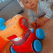 Lynao a rejoint le concours — aidez-le/la à gagner de superbes lots ! baby, blue_wheel, child, colorful_toy, couch, curious, face, floor, floor_play, indoor, living_room, pajamas, person, play_area, red_toy, striped_clothing, toy, wheel, wooden_floor, young_child