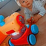 baby, child, toy, floor, wooden_floor, pajamas, indoor, living_room, couch, play_area, wheel, colorful_toy, person, face, young_child, curious, floor_play, blue_wheel, red_toy, striped_clothing
