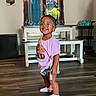 balloons, cabinet, child, colorful_socks, denim_shorts, furniture, girl, glasses, graphic_tshirt, happy, home, indoor, living_room, mirror, portrait, purple_shirt, smiling, standing, toddler, wooden_floor