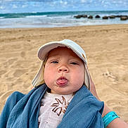 Luka is registered to the contest to win money with this photo: baby, child, face, tongue, hat, blue_blanket, beach, sand, ocean, sky, clouds, person, cute, infant, summer, outdoor, playful, portrait, headwear, expression
