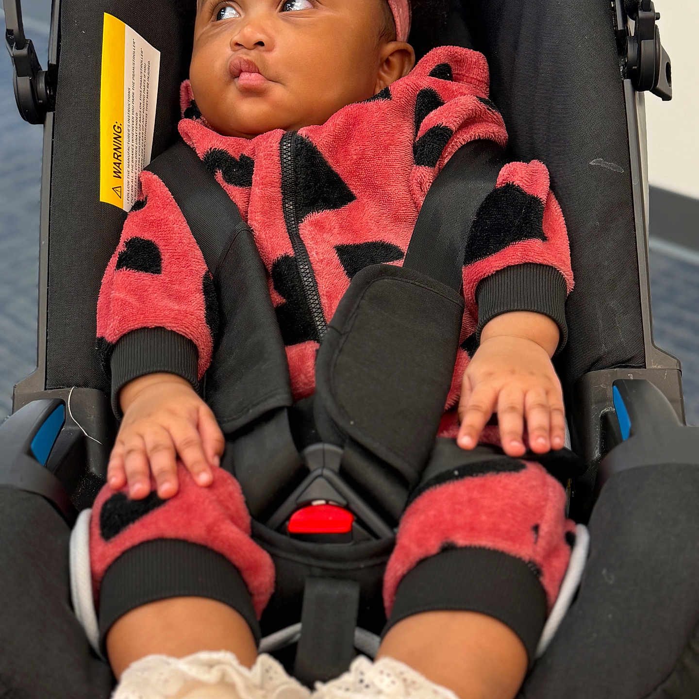 Rayanne is registered to the contest to win money with this photo: baby, child, car_seat, seatbelt, pink_headband, fuzzy_clothing, red_clothing, black_clothing, socks, white_socks, lace_trim, indoor, portrait, infant, cute, pouty_expression, curious, hands, feet, person
