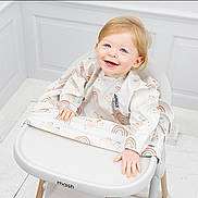 Angelica Emery is registered to the contest to win money with this photo: toddler, child, smiling, high_chair, bib, rainbow_pattern, unicorn_pattern, blonde_hair, happy, indoor, white_floor, white_wall, wooden_legs, modern_furniture, cute, baby, portrait, sitting, playful, bright