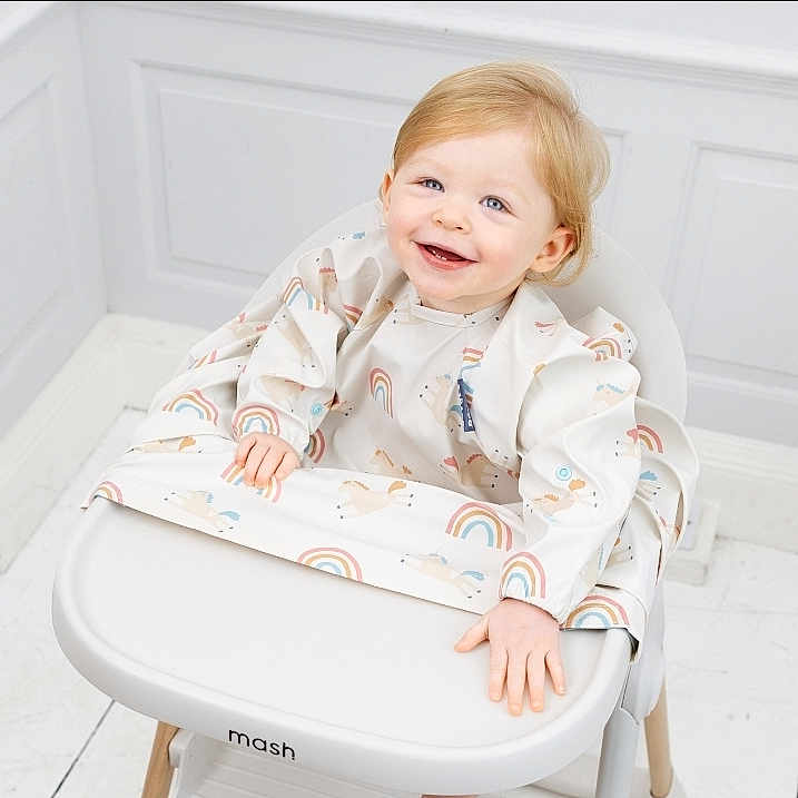Angelica Emery is registered to the contest to win money with this photo: baby, bib, blonde_hair, bright, child, cute, happy, high_chair, indoor, modern_furniture, playful, portrait, rainbow_pattern, sitting, smiling, toddler, unicorn_pattern, white_floor, white_wall, wooden_legs