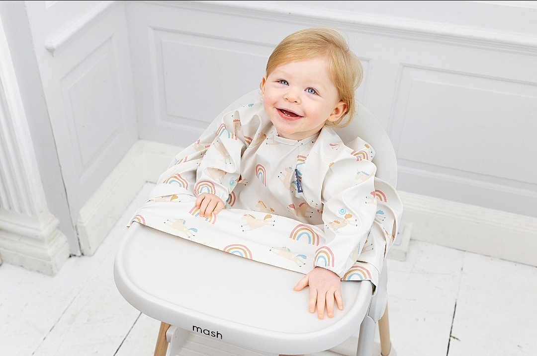 Angelica Emery is registered to the contest to win money with this photo: toddler, child, smiling, high_chair, bib, rainbow_pattern, unicorn_pattern, blonde_hair, happy, indoor, white_floor, white_wall, wooden_legs, modern_furniture, cute, baby, portrait, sitting, playful, bright