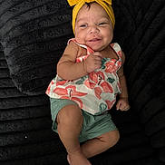 Marisol is registered to the contest to win money with this photo: baby, child, smile, headband, yellow, floral, top, green_shorts, barefoot, black_blanket, texture, cute, happy, infant, person, indoors, portrait, skin, small_hand, feet