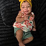 baby, child, smile, headband, yellow, floral, top, green_shorts, barefoot, black_blanket, texture, cute, happy, infant, person, indoors, portrait, skin, small_hand, feet