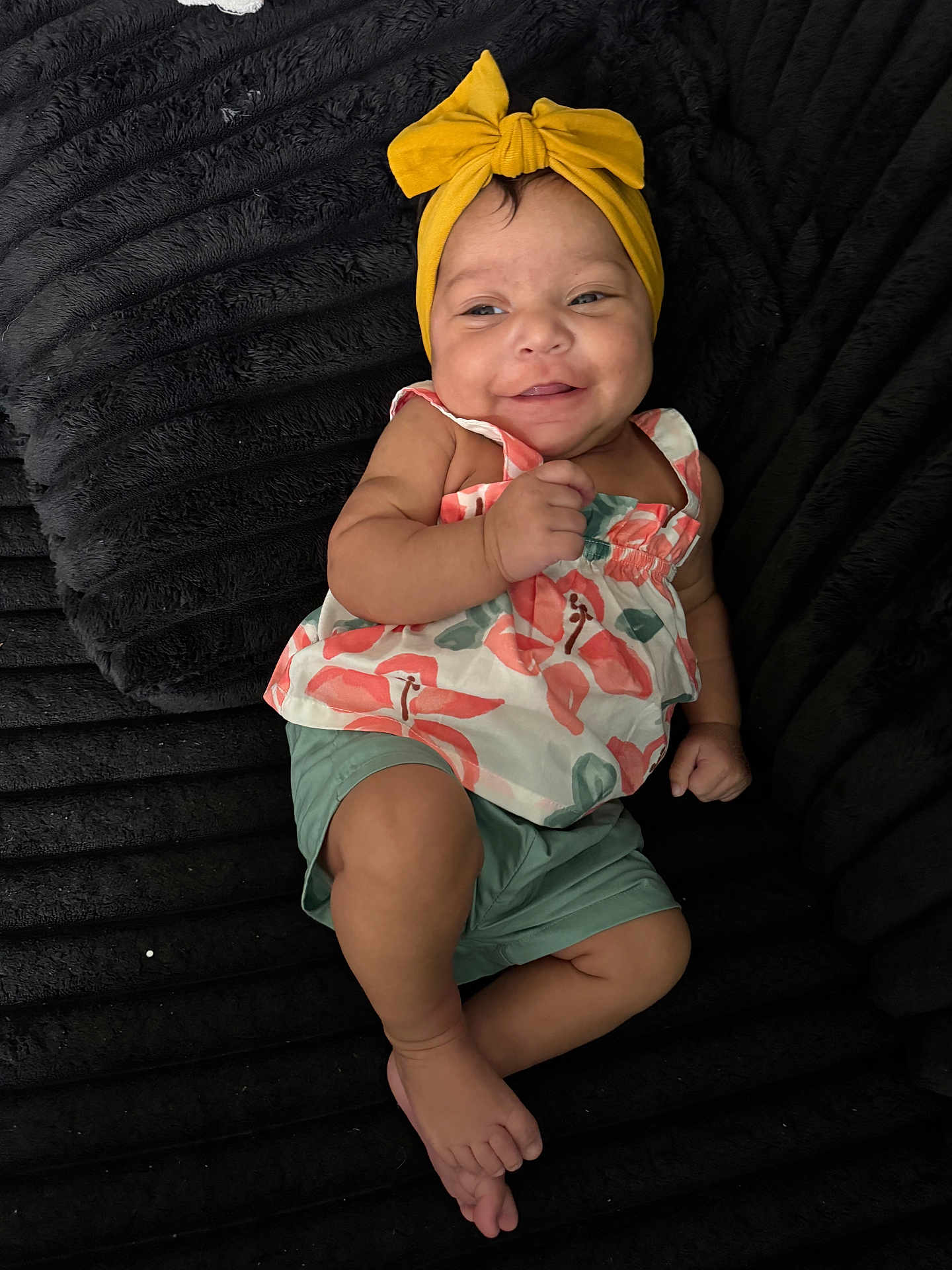 Marisol is registered to the contest to win money with this photo: baby, child, smile, headband, yellow, floral, top, green_shorts, barefoot, black_blanket, texture, cute, happy, infant, person, indoors, portrait, skin, small_hand, feet