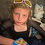 child, blonde_hair, sunglasses, chips, couch, pillow, casual_clothing, indoor, snack, blue_bag, person, young, sitting, face, looking_at_camera, hand, relaxed, home, comfortable, expression