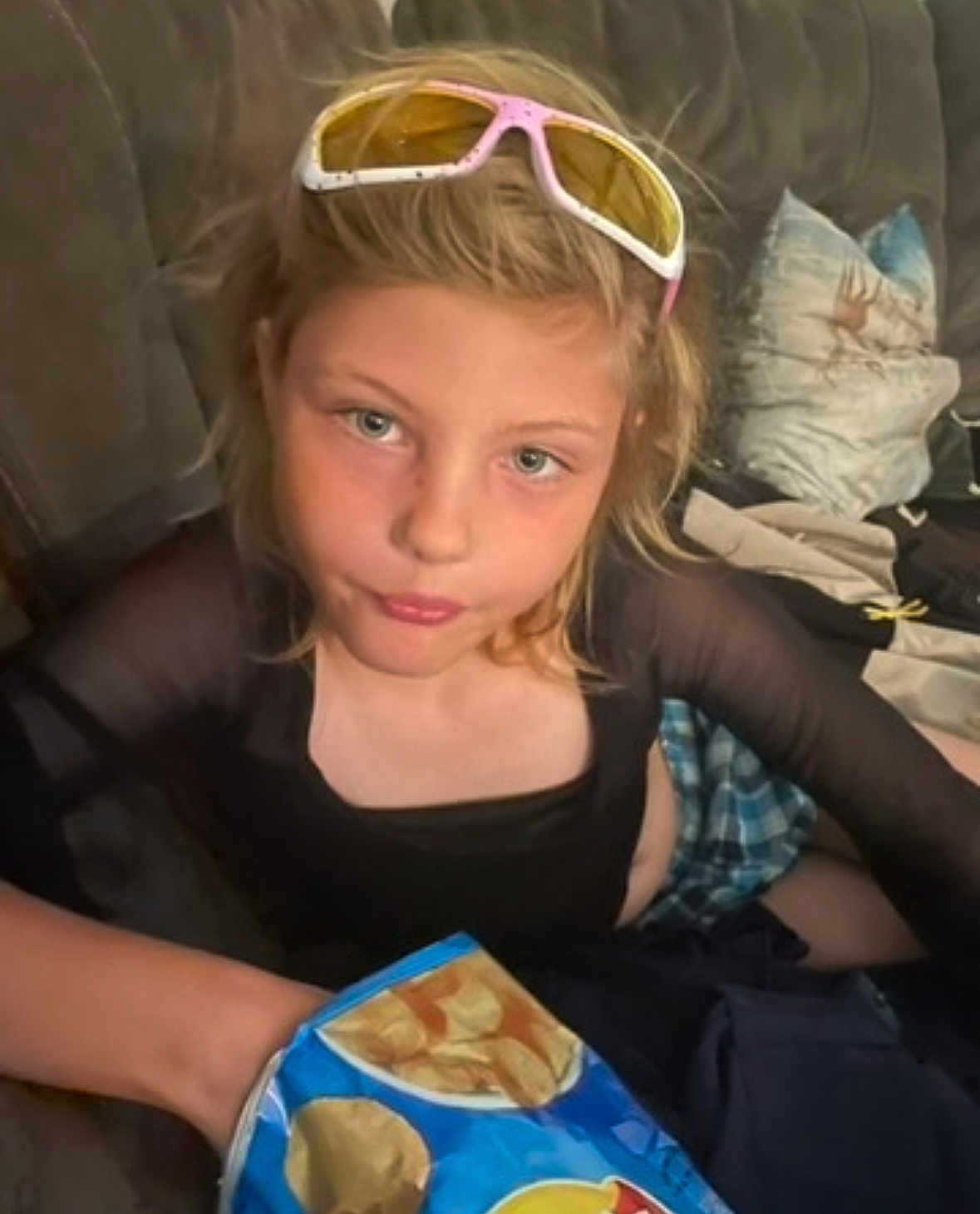 Gracelyn joined the competition — help win amazing prizes! child, blonde_hair, sunglasses, chips, couch, pillow, casual_clothing, indoor, snack, blue_bag, person, young, sitting, face, looking_at_camera, hand, relaxed, home, comfortable, expression