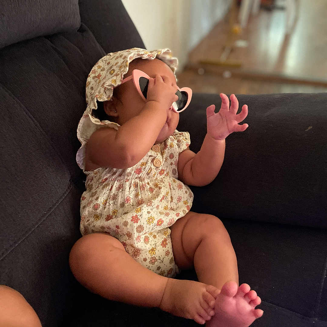 Anyla is registered to the contest to win money with this photo: adorable, baby, casual, child, comfortable, couch, floral_clothing, foot, furniture, hand, hat, home, indoor, infant, person, playful, relaxed, seated, skin, sunglasses