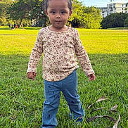 Salome is registered to the contest to win money with this photo: child, toddler, girl, portrait, outdoor, park, grass, lawn, standing, face, hair, pigtails, floral_shirt, blue_pants, serious_expression, sunlight, trees, building, people, nature