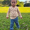 child, toddler, girl, portrait, outdoor, park, grass, lawn, standing, face, hair, pigtails, floral_shirt, blue_pants, serious_expression, sunlight, trees, building, people, nature
