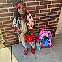 child, girl, brick_wall, backpack, cartoon_character, toy, red_shoes, white_tights, strawberry_pattern, hair_beads, pink_bow, sidewalk, outdoor, smiling, posing, clothing, accessory, happy, person, casual