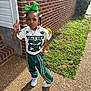 Chozyn is registered to the contest to win money with this photo: child, green_bow, athletic_wear, white_shoes, brick_wall, sidewalk, grass, outdoor, sunlight, pose, hand_gesture, smile, person, young, casual_clothing, hair_beads, necklace, shadow, window, building