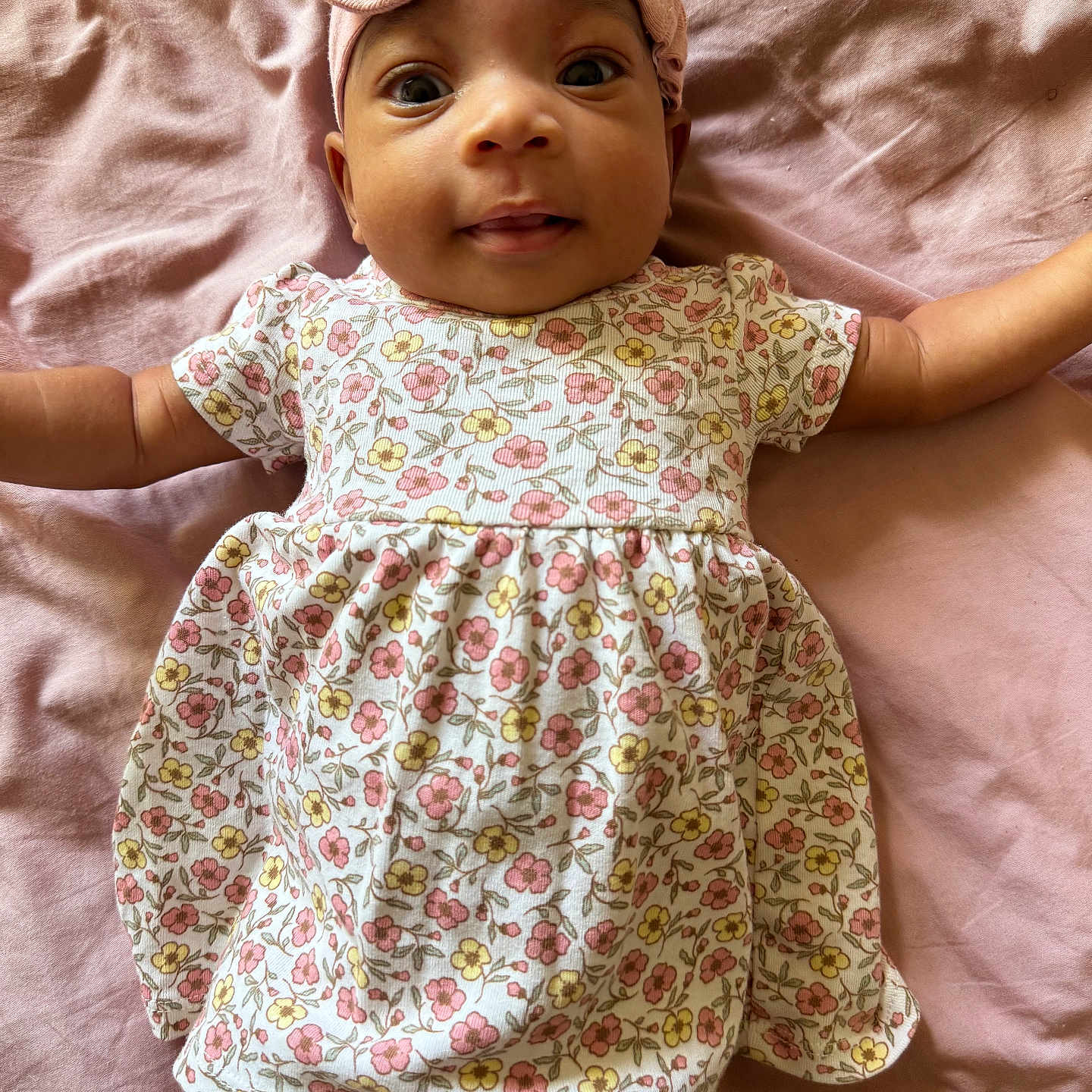 Diyonne is registered to the contest to win money with this photo: baby, blanket, child, close_up, cute, face, floral_dress, happy, headband, indoors, infant, lying_down, one_person, outstretched_arms, pink_bow, portrait, skin, smiling, soft_fabric, wide_eyes