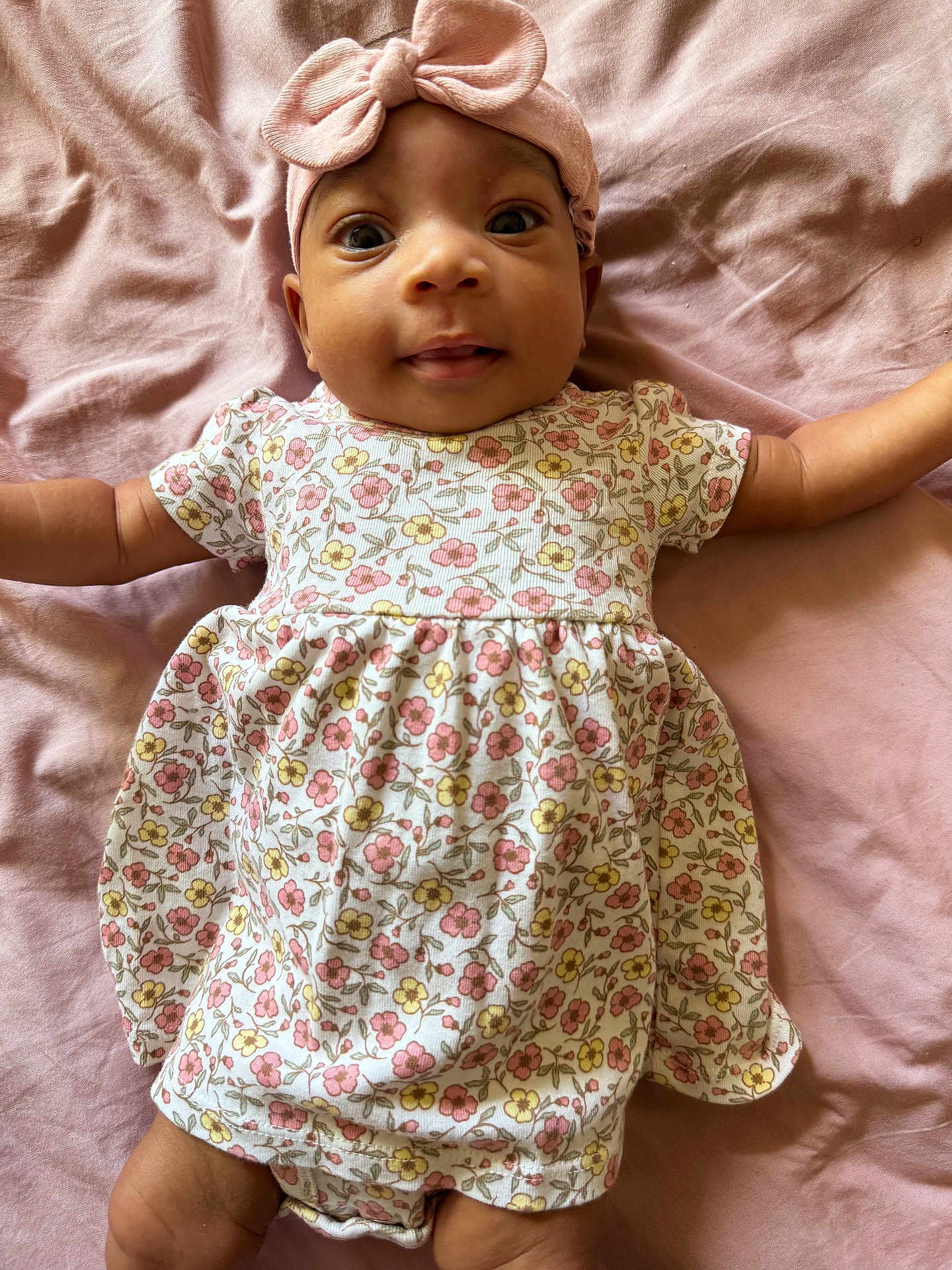 Diyonne is registered to the contest to win money with this photo: baby, infant, child, pink_bow, headband, floral_dress, blanket, soft_fabric, lying_down, cute, wide_eyes, outstretched_arms, portrait, smiling, happy, indoors, one_person, close_up, skin, face