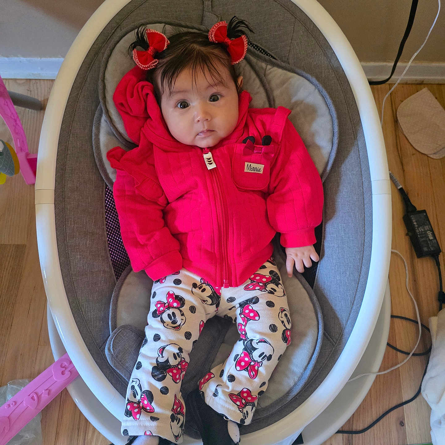 Luxana is registered to the contest to win money with this photo: baby, baby_rocker, black_shoes, child, clothing, cute, expression, face, floor_mat, hair_bows, home, indoor, infant, minnie_mouse, pants, person, red_jacket, seated, toy, wood_floor