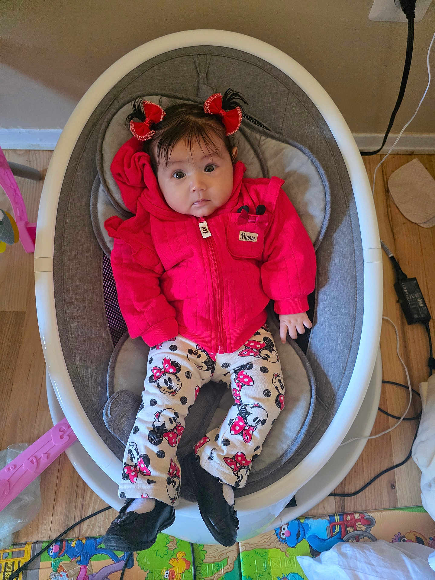 Luxana is registered to the contest to win money with this photo: baby, infant, child, red_jacket, minnie_mouse, pants, black_shoes, baby_rocker, floor_mat, wood_floor, indoor, cute, clothing, person, face, home, toy, hair_bows, expression, seated