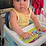 baby, child, high_chair, yellow_clothing, rainbow_pattern, indoor, furniture, tray, hand, face, curious, black_hair, crib, red_clothing, carpet, earrings, window, home, person, seated