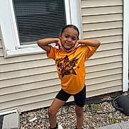 Truu is registered to the contest to win money with this photo: child, posing, yellow_shirt, black_shorts, white_shoes, outdoor, window, house_siding, stones, playful, smiling, young, person, footwear, sidewalk, casual, happy, standing, daylight, fashion
