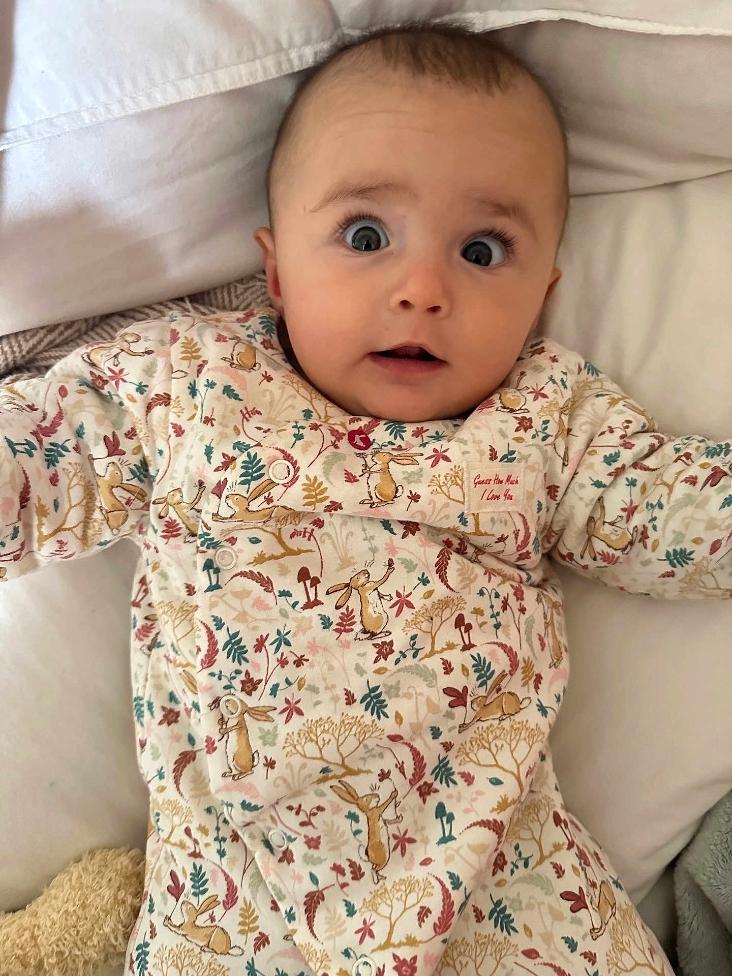 Ella joined the competition — help win amazing prizes! baby, infant, onesie, patterned_clothing, bed, blanket, curious_expression, wide_eyes, soft_lighting, indoor, person, cute, child, face, head, plush, comfort, sleepwear, lying_down, surprise