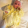 Annie joined the competition — help win amazing prizes! baby, pacifier, headband, bow, yellow_sweater, knitwear, onesie, infant, bed, pillow, sleepwear, hand, face, cute, child, newborn, soft_lighting, indoor, portrait, lying_down