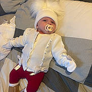 Annie is registered to the contest to win money with this photo: baby, pacifier, hat, white_sweater, red_pants, boots, bed, blanket, striped, lying_down, infant, cute, warm_clothing, gloves, indoor, child, face, portrait, soft_toys, resting