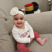 Tia is registered to the contest to win money with this photo: baby, child, smiling, headband, flower, red_roses, striped_pants, couch, indoor, table, box, white_shirt, happy, person, portrait, sitting, cozy, cute, infant, home