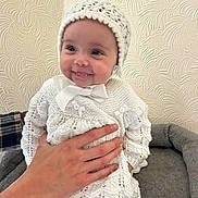 Lola joined the competition — help win amazing prizes! baby, smile, hand, knitwear, bonnet, white_clothing, infant, cute, portrait, indoors, wall_texture, soft_cushion, child, happy, person, sitting, cozy, young, warm, hands