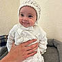 baby, smile, hand, knitwear, bonnet, white_clothing, infant, cute, portrait, indoors, wall_texture, soft_cushion, child, happy, person, sitting, cozy, young, warm, hands