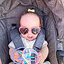 baby, child, sunglasses, heart_shaped, toy, stroller, gray, yellow_dress, hair_bow, cute, infant, outdoor, sunny, smiling, hands, face, seatbelt, playful, portrait, person